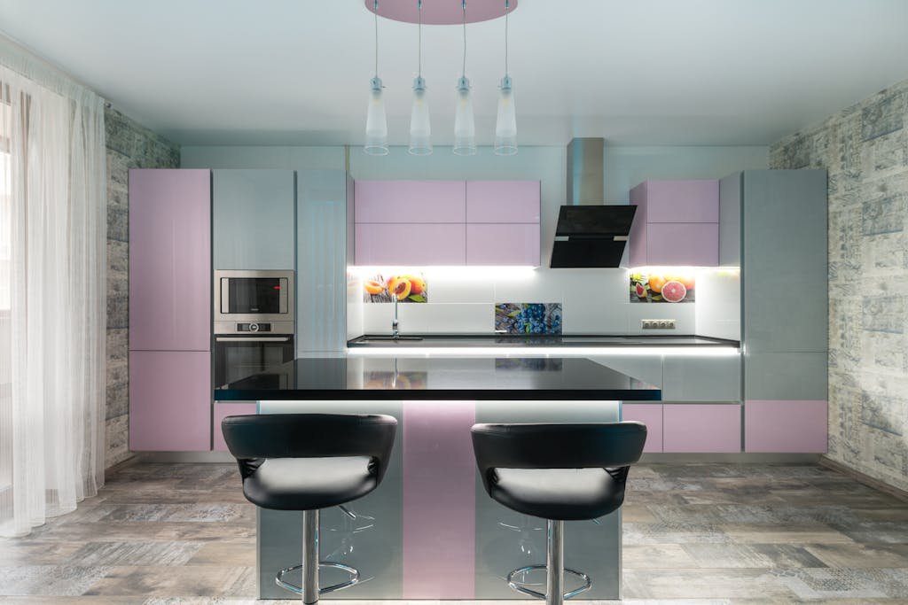Modern kitchen interior with pastel pink and gray cabinets, island counter, and bar stools under pendant lights.