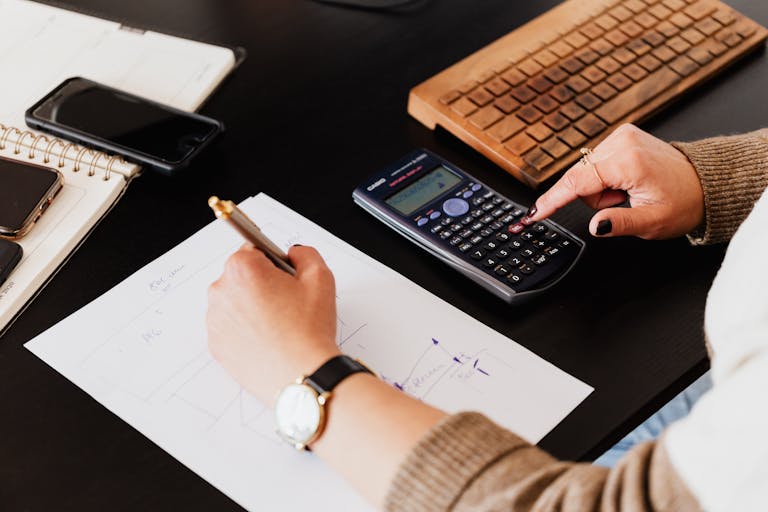 Close-up of hands working with a calculator and notebook on a desk, analysing documents.