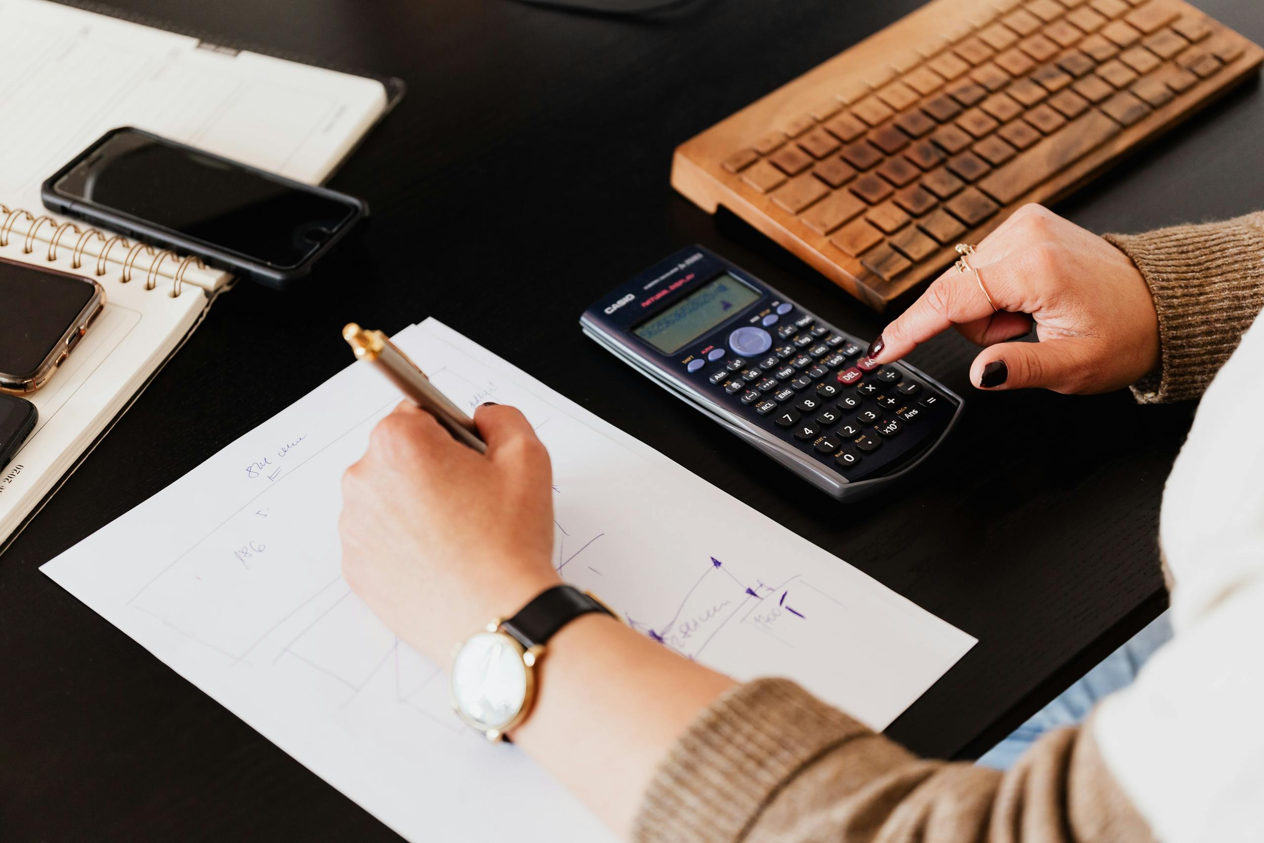 Close-up of hands working with a calculator and notebook on a desk, analysing documents.